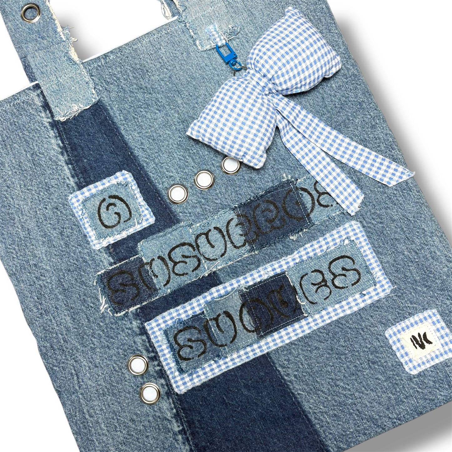Close-up of the denim patchwork, embroidered text, and blue gingham bow charm on the A Susurros Suaves Tote bag.