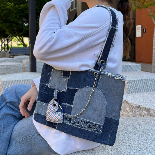 Woman wearing white sweatshirt and jeans while holding the Blue Denim Handbag, seated outdoors in an urban setting.