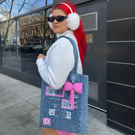 Street-style shot of a model in sunglasses and earmuffs wearing the Cada Vez Que Me Miras tote bag on her shoulder, showing off the bag's stitched text and pink bow.