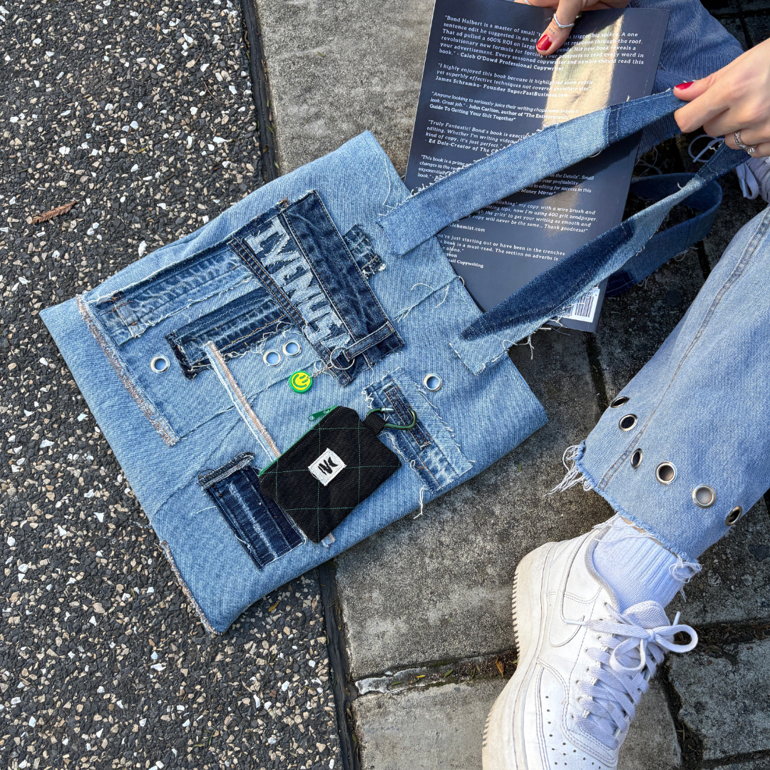 Flat lay of the Iconic Green tote bag placed on pavement with a book and worn by a person in denim pants and white sneakers.