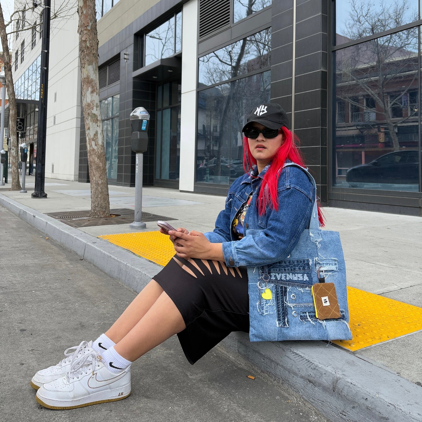 Woman sitting on a sidewalk wearing a black skirt and denim jacket, holding the Iconic Yellow tote bag with a yellow heart charm and brown pouch.