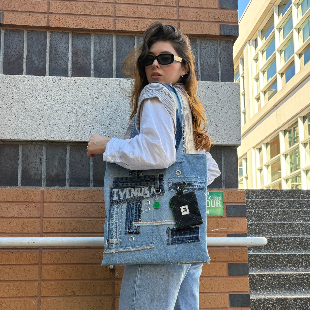 Woman in sunglasses and streetwear holding the Iconic Green denim tote against a modern brick building.