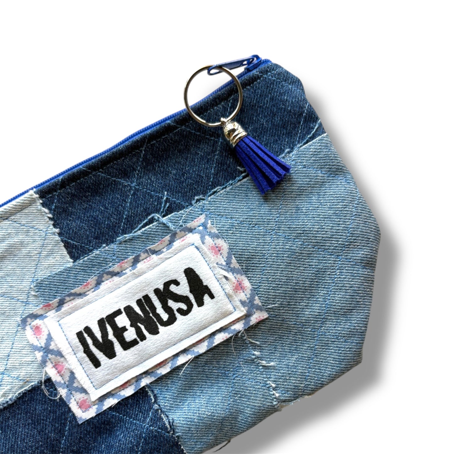 Close-up of IVENUSA patchwork denim makeup bag showing stitched logo, blue tassel zipper pull, and quilted texture