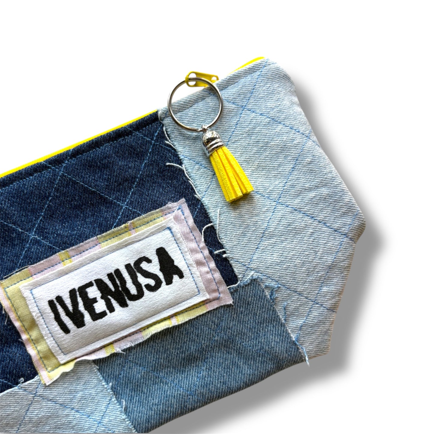 Close-up of IVENUSA patchwork denim makeup bag showing stitched logo, yellow tassel zipper pull, and quilted texture