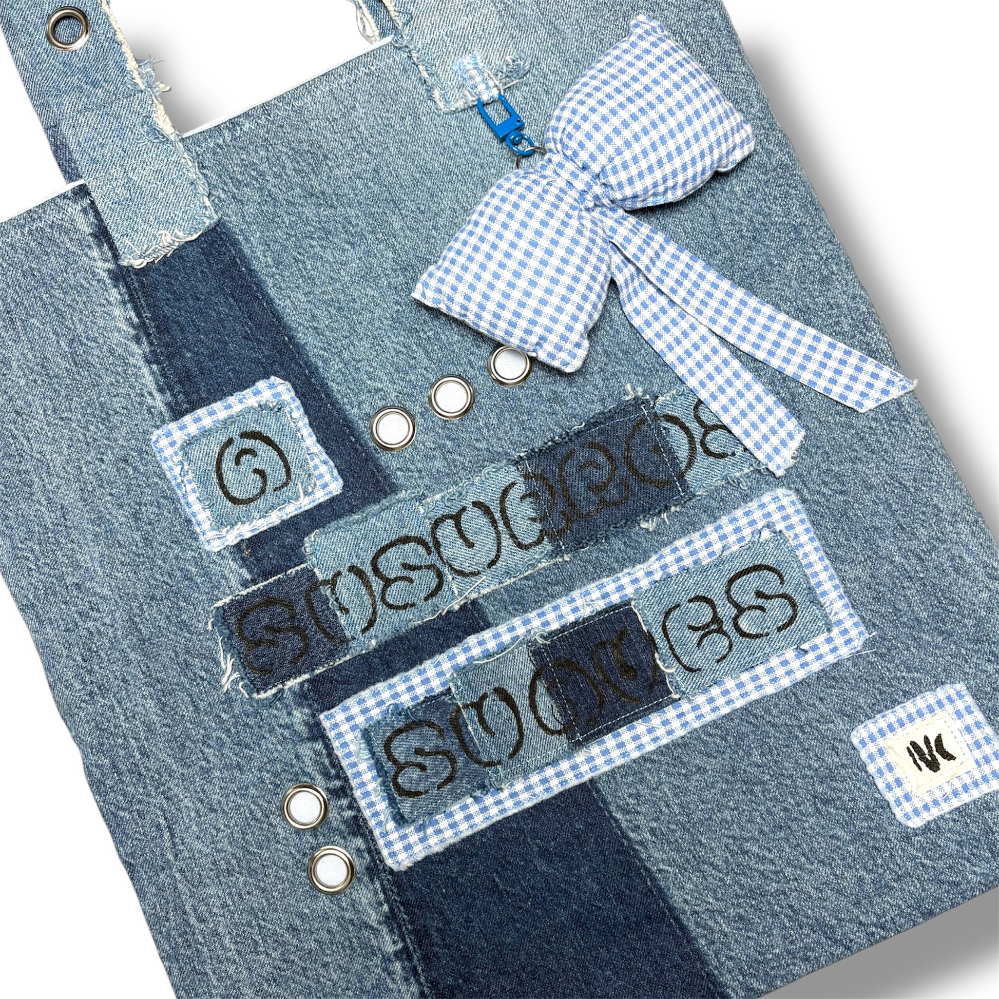 Close-up of the denim patchwork, embroidered text, and blue gingham bow charm on the A Susurros Suaves Tote bag.