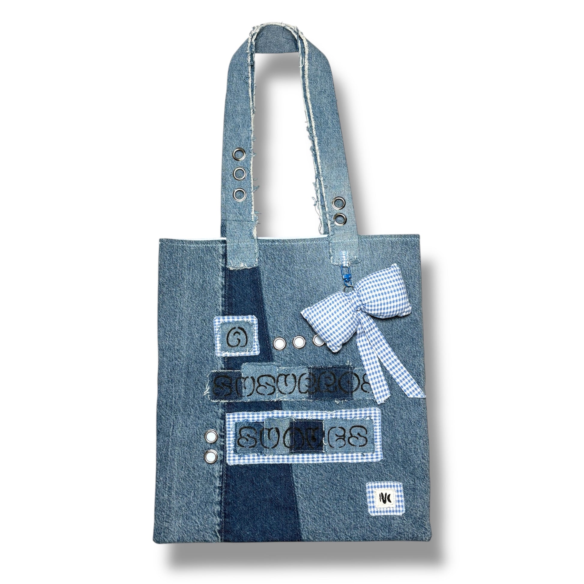  Front view of the A Susurros Suaves Tote bag, featuring patchwork denim with grommets and a blue gingham bow charm.