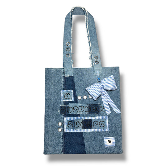  Front view of the A Susurros Suaves Tote bag, featuring patchwork denim with grommets and a blue gingham bow charm.