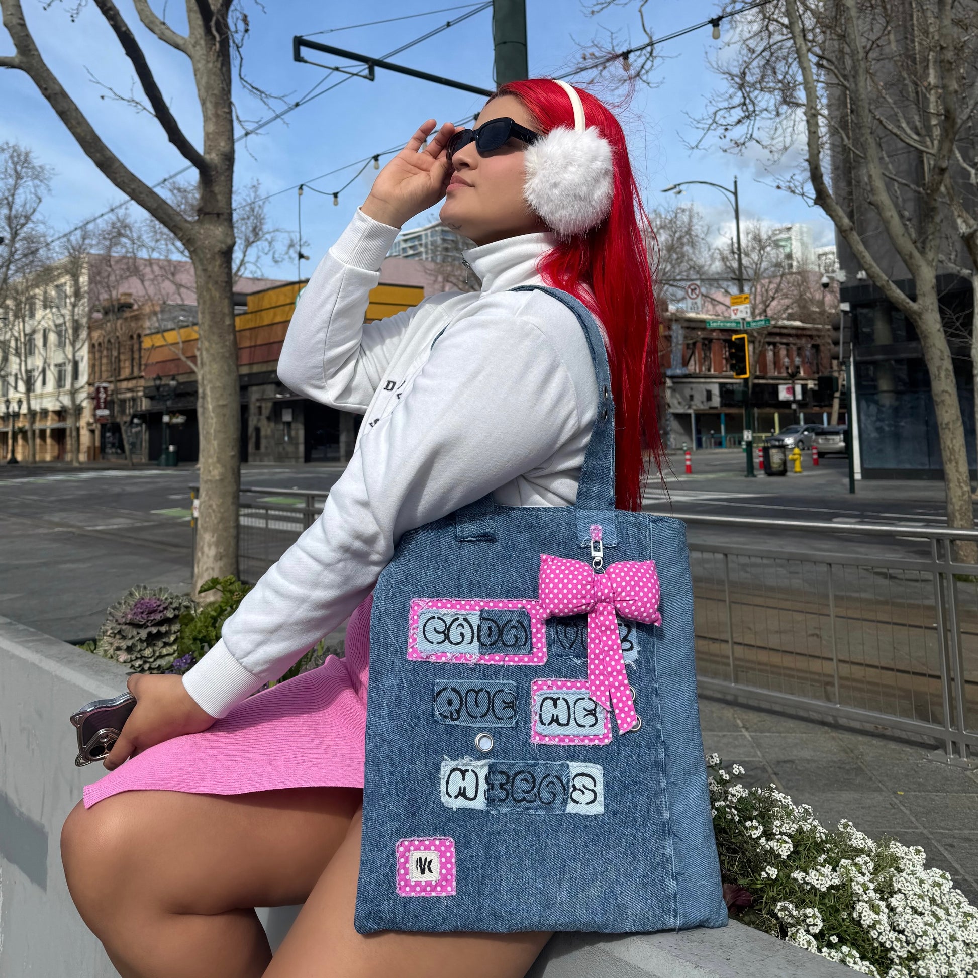 Model with bright red hair, white earmuffs, and a white turtleneck sitting on a ledge in the city, styling the Cada Vez Que Me Miras denim tote with a pink polka dot bow.