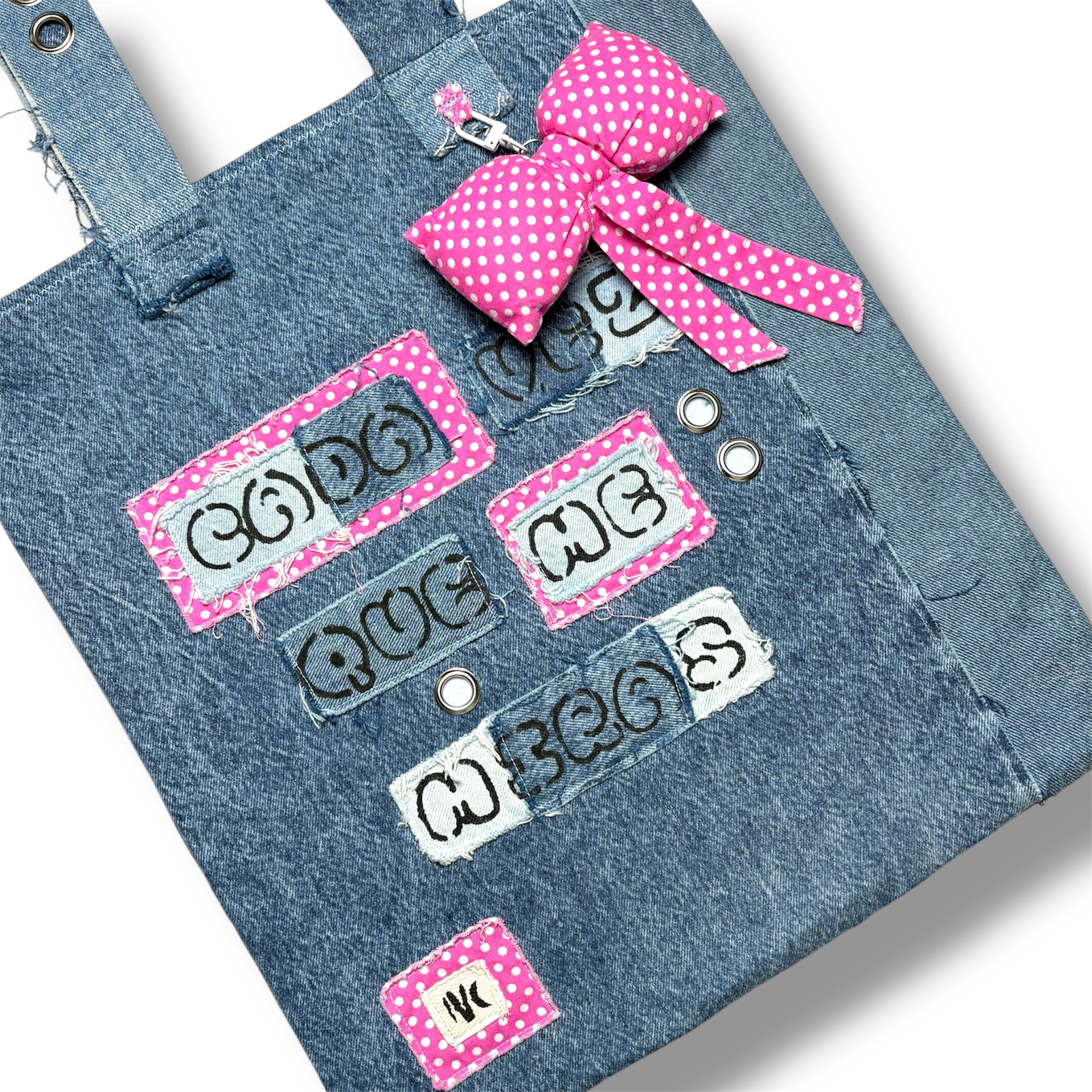 Close-up of the Cada Vez Que Me Miras tote showing embroidered words and stitched denim panels with pink accents and grommet detailing.