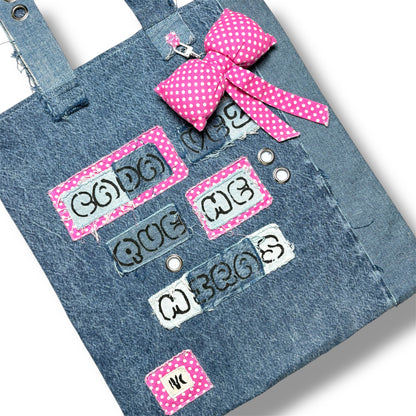 Close-up of the Cada Vez Que Me Miras tote showing embroidered words and stitched denim panels with pink accents and grommet detailing.