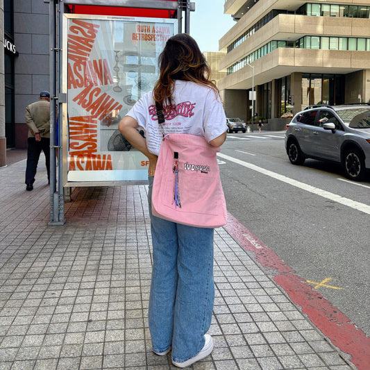 Woman wearing jeans and a graphic t-shirt carrying a pink IVENUSA Design shoulder bag with colorful tassel keychain on a city street.