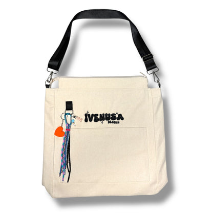 IVENUSA Rush Hour Bag in neutral beige with front pocket, black embroidered logo, and detachable orange heart charm with multicolor tassels.