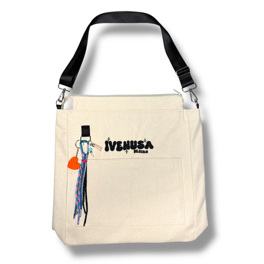IVENUSA Rush Hour Bag in neutral beige with front pocket, black embroidered logo, and detachable orange heart charm with multicolor tassels.
