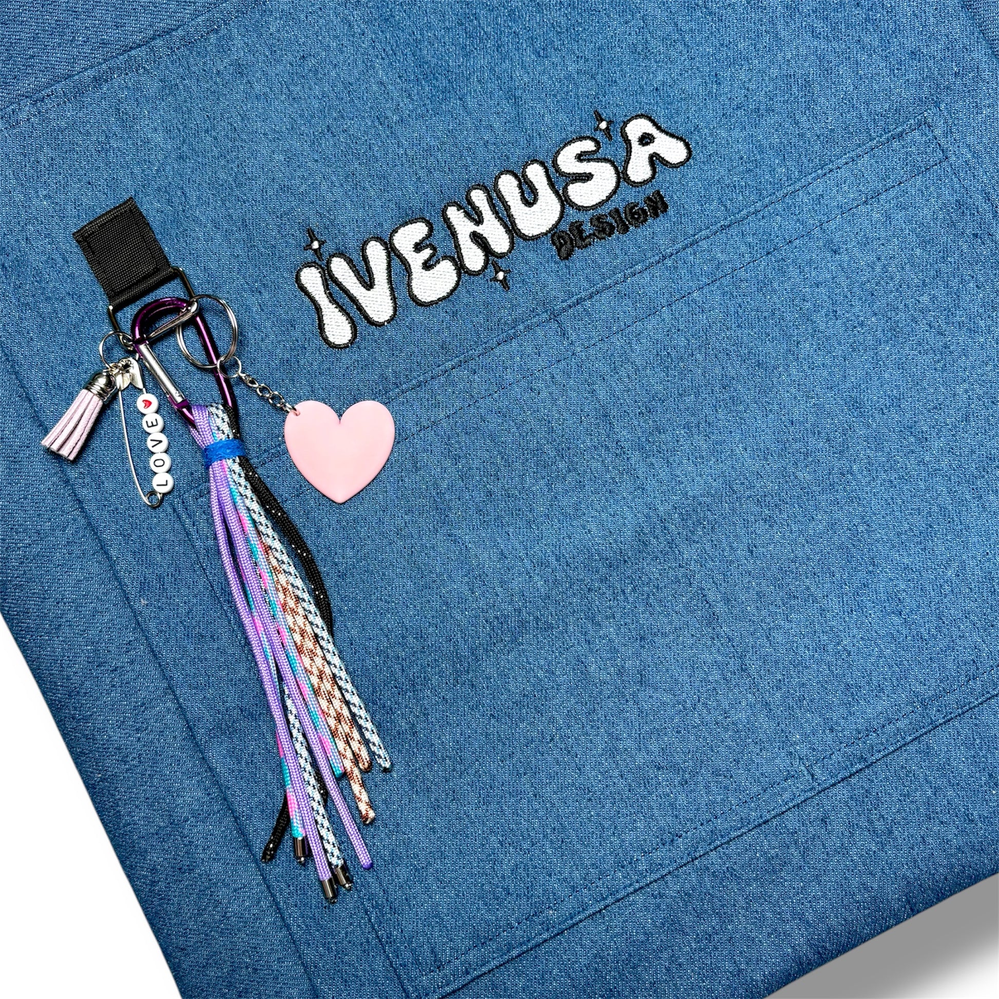Close-up of the Skyline Bag’s IVENUSA embroidery with pastel accessories including a pink heart charm and braided tassel keychain set.