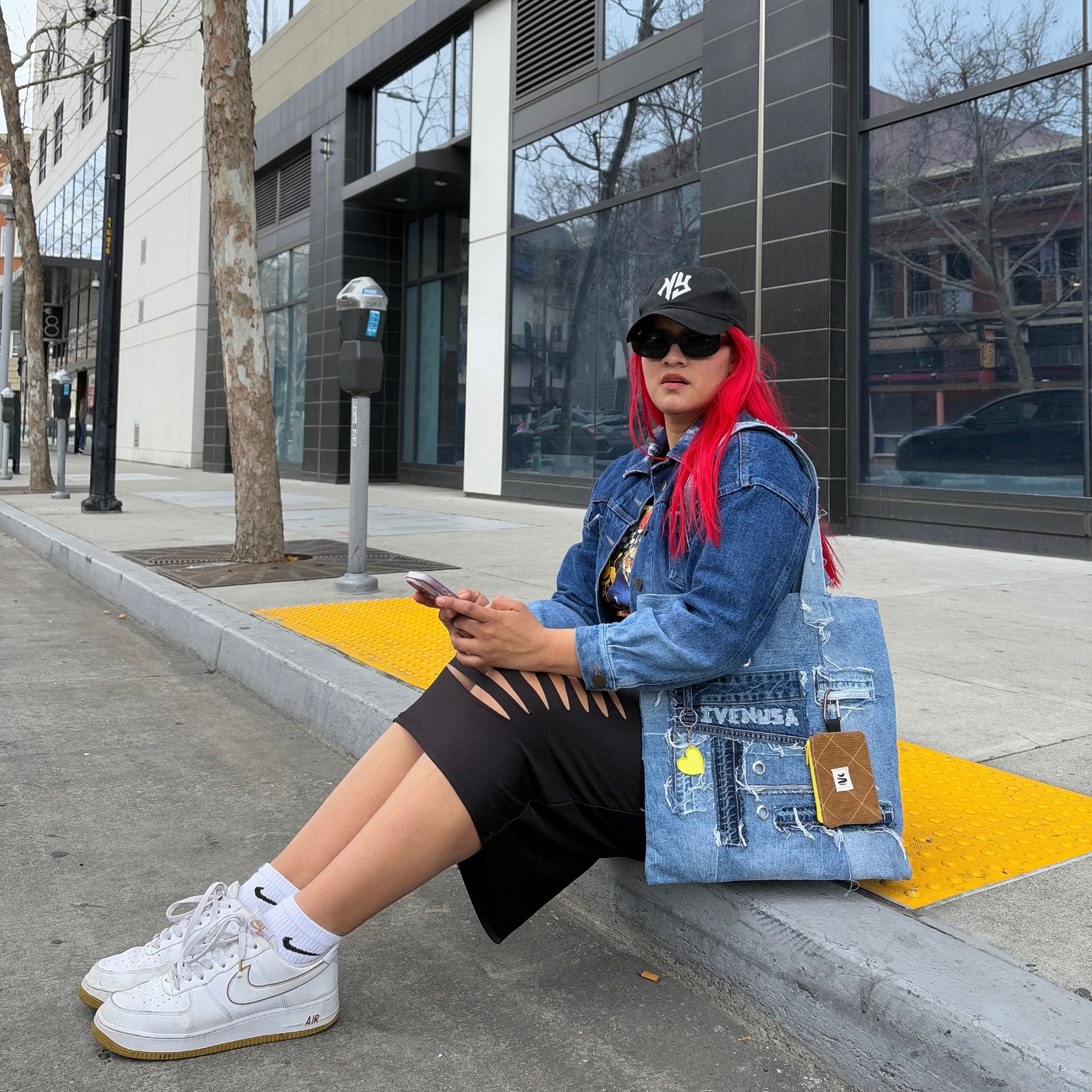 Woman sitting on a sidewalk wearing a black skirt and denim jacket, holding the Iconic Yellow tote bag with a yellow heart charm and brown pouch.