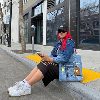 Woman sitting on a sidewalk wearing a black skirt and denim jacket, holding the Iconic Yellow tote bag with a yellow heart charm and brown pouch.