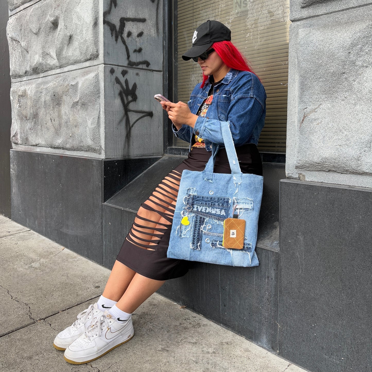Model seated by a city wall, styling the Iconic Yellow denim tote with visible heart keychain and utility pouch.