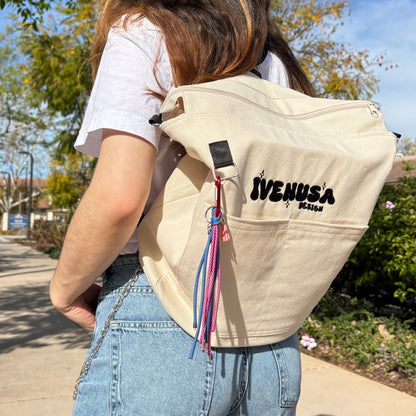 Model wearing the Rush Hour Bag as a backpack on a sunny day, showcasing the beige canvas material and playful keychain charms with pink and blue rope tassels.