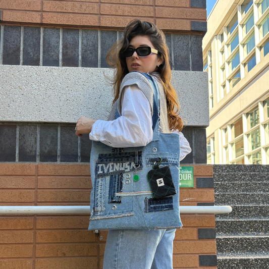 Woman in sunglasses and streetwear holding the Iconic Green denim tote against a modern brick building.