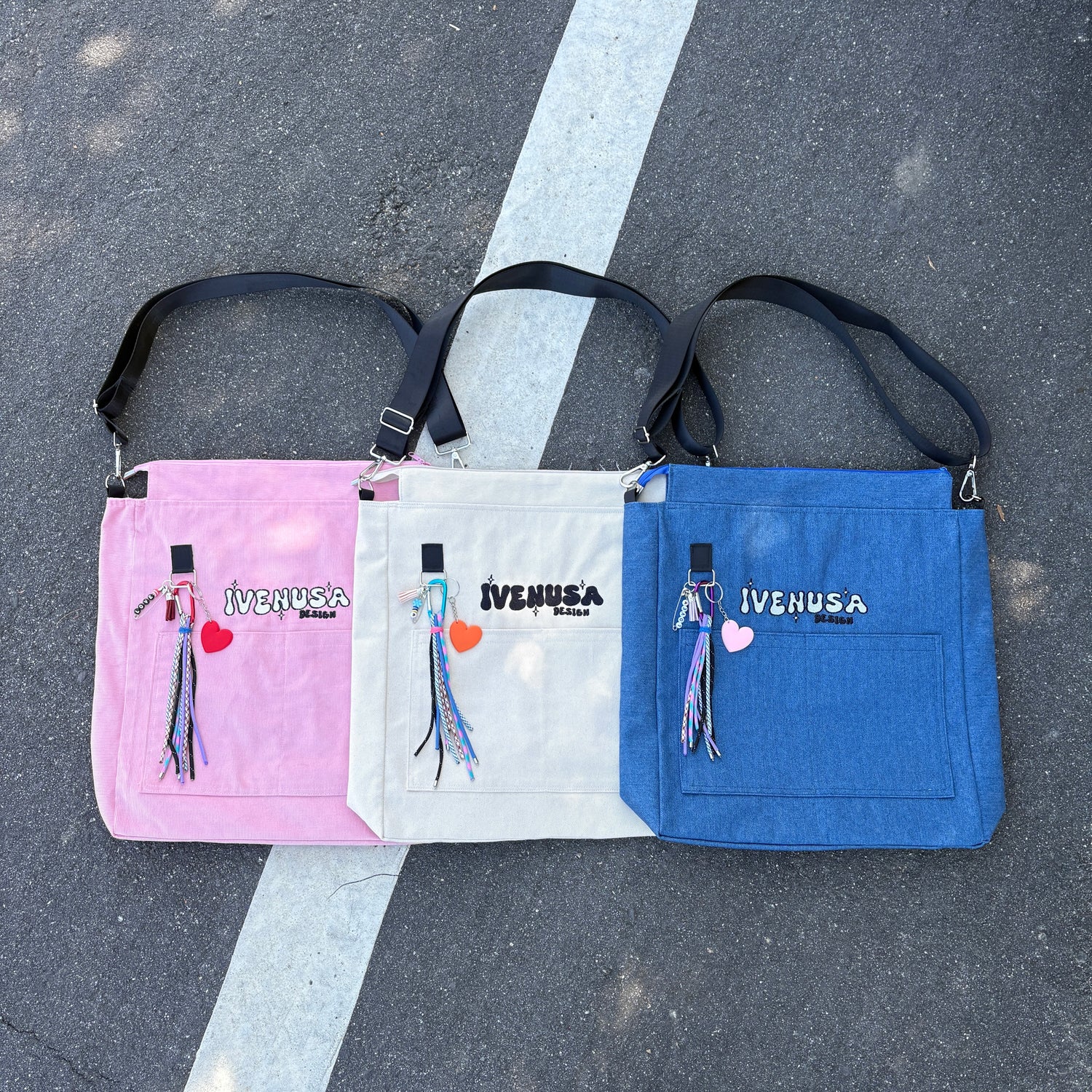 Urban Nomad Collection tote bags by IVENUSA in pink, cream, and denim blue, each featuring embroidered logos, fringe keychains, and adjustable black straps, displayed side by side on asphalt.