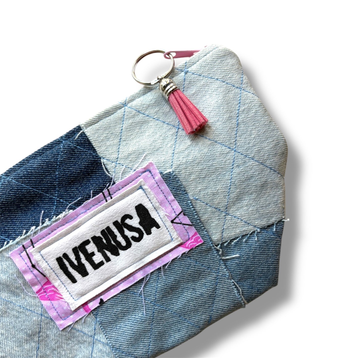Close-up of IVENUSA patchwork denim makeup bag showing stitched logo, blush tassel zipper pull, and quilted texture